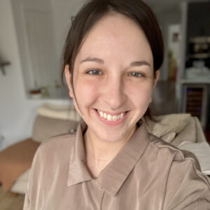 Close-up of Corrine Lajoie smiling. They are a white person with dark brown hair and green eyes, wearing a beige silky button-up and a small nose ring. Background is blurred.